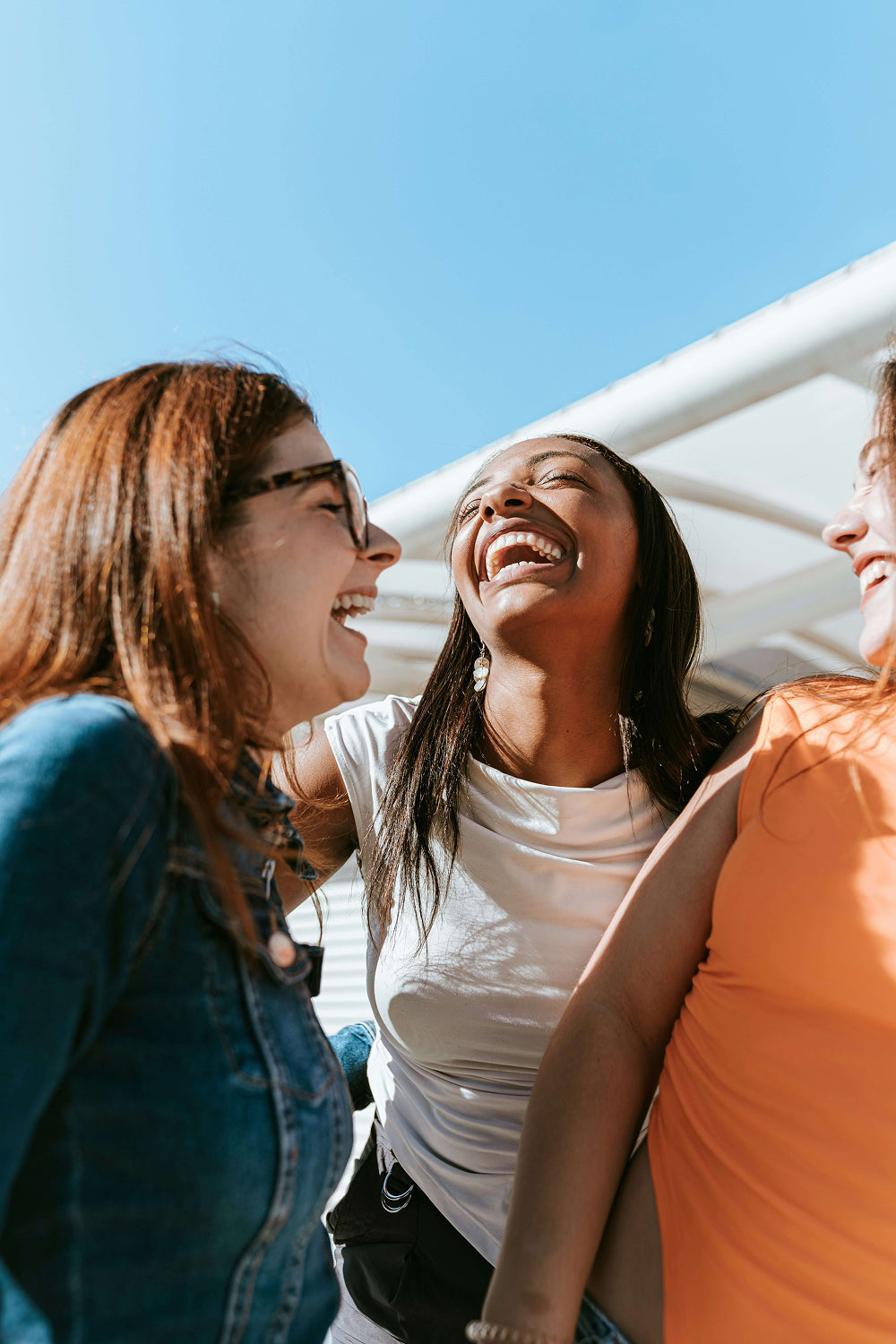 Three women laughing together outdoors under a clear blue sky.